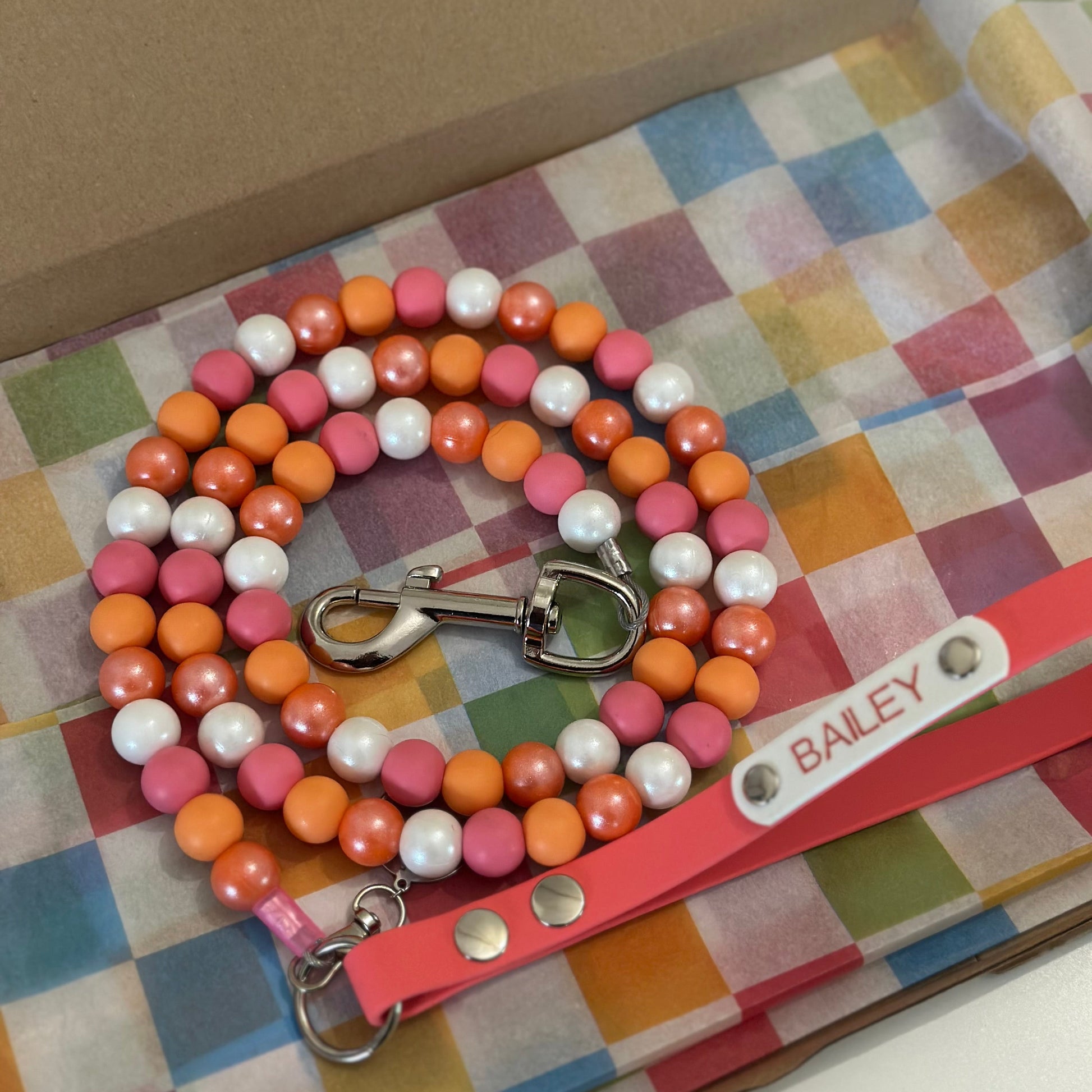A pink leather pet leash with silver hardware and a white name tag reading 'BAILEY' is displayed on a colorful checkered tissue paper inside a brown cardboard box. The leash is paired with a matching beaded collar made of pink, orange, and white beads.
