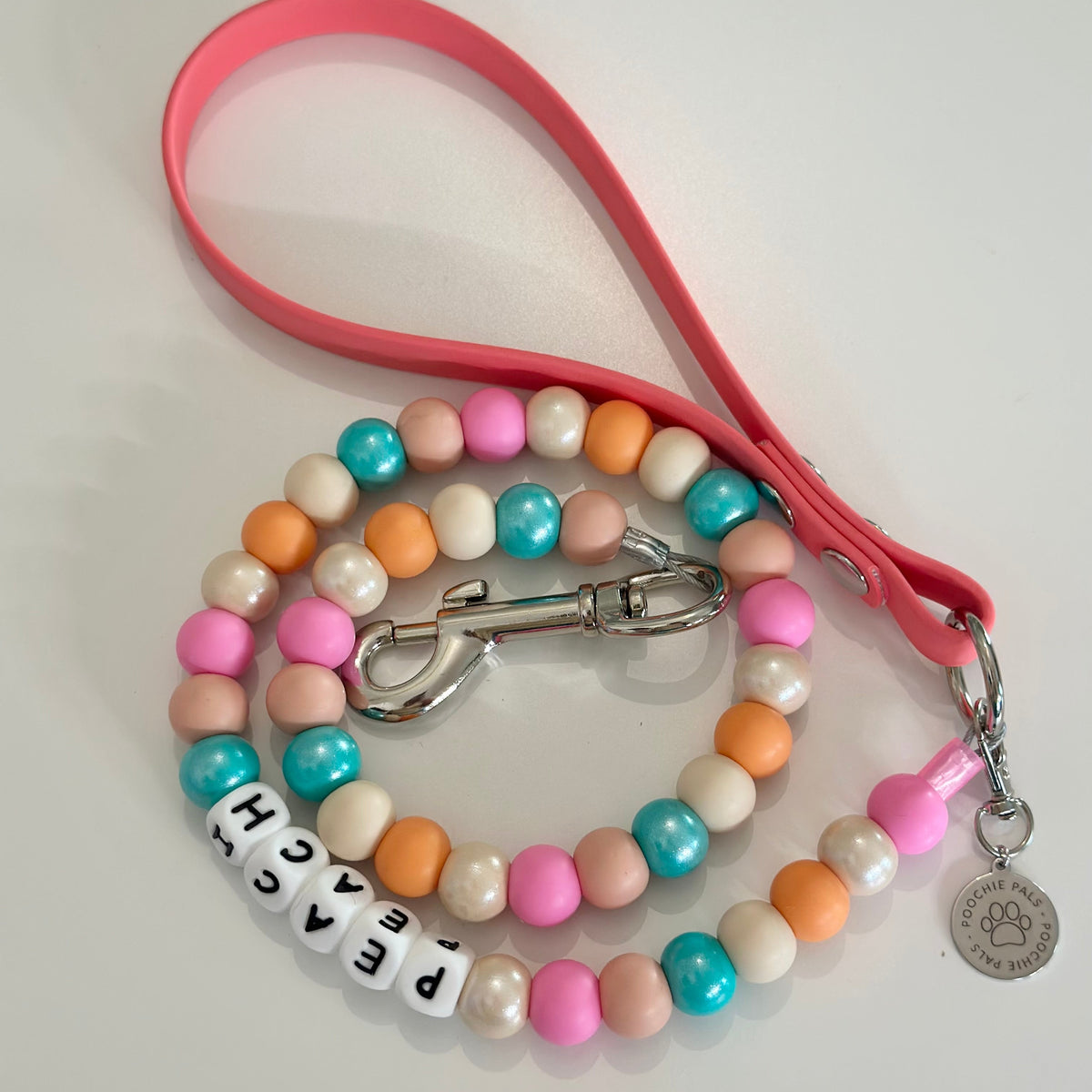 A beaded dog lead with a matching beaded strap coiled next to it on a white surface. The beaded strap features pastel pink, orange, teal blue, and cream beads, with white letter beads spelling 'PEACH' and a silver metal clasp and charm.