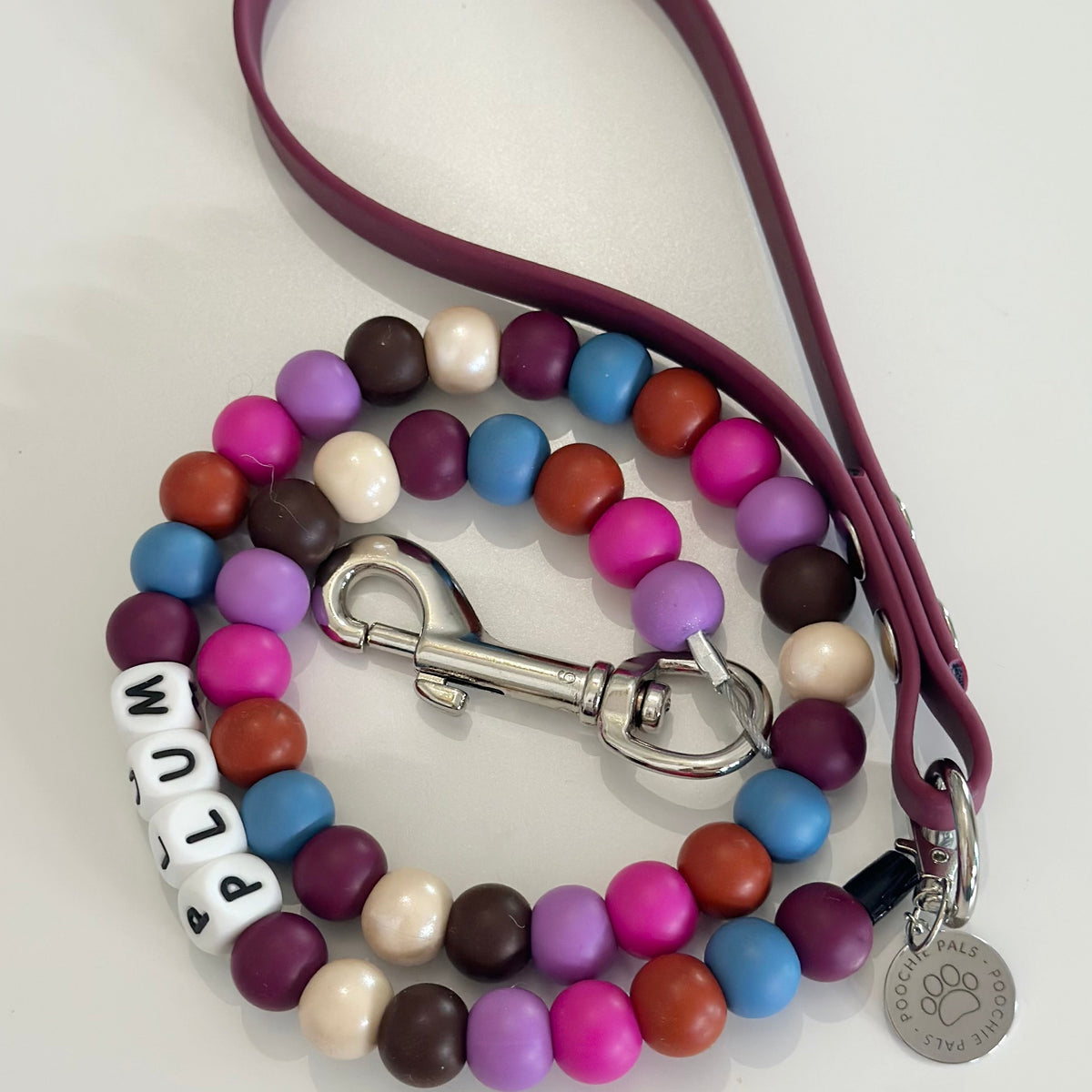 A beaded dog lead featuring a dark purple biothane strap and a matching beaded collar. The collar is decorated with colorful round beads in pink, purple, blue, orange, brown, and cream, accented by white letter beads spelling 'PLUM' and a silver metal clasp and paw-print tag.