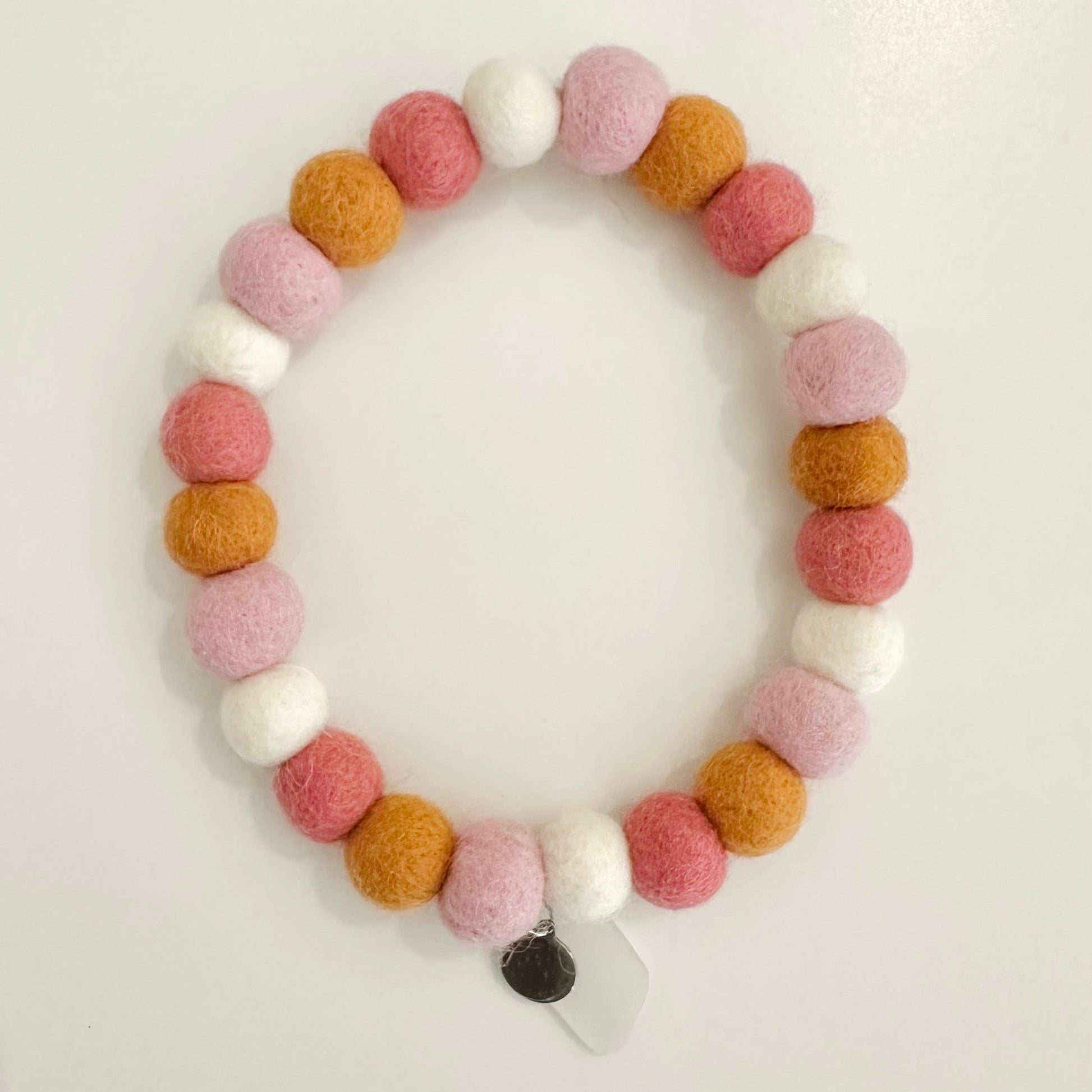 A beaded stretch bracelet made of alternating felt pom-poms in dusty pink, mustard yellow, light pink, and white, with a small white tag attached at the bottom, displayed on a plain white surface. featuring a Cornish sea glass charm