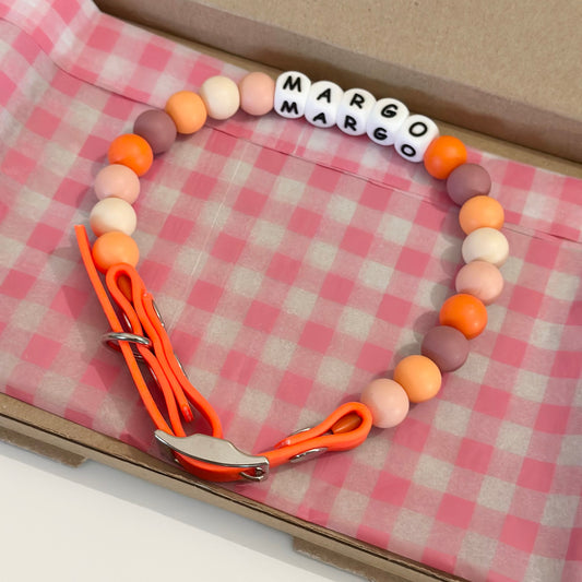 A beaded dog collar with orange, peach, pink, and white beads, featuring a white letter bead spelling 'MARGO' and bright orange biothane strap with a silver clasp, displayed inside a brown box lined with a pink and white checkered paper.