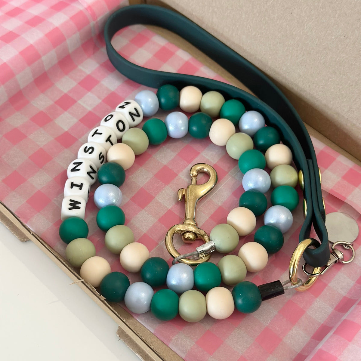 A beaded dog leash with a dark green strap and matching green and cream beads, displayed inside a brown cardboard box lined with pink gingham paper. The leash features a gold-tone clasp and a white bead section spelling 'WINSTON'.