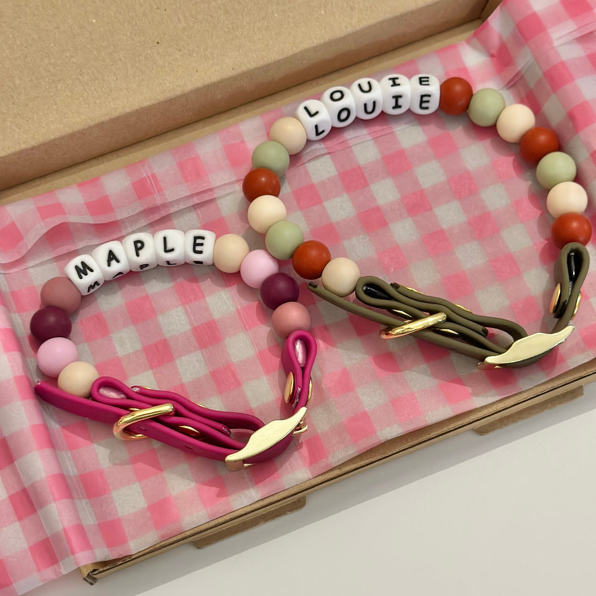 Two beaded dog collars displayed inside an open kraft gift box lined with pink-and-white gingham paper. One dog collar is labeled 'MAPLE' in white letter beads and features a magenta cord with gold hardware.