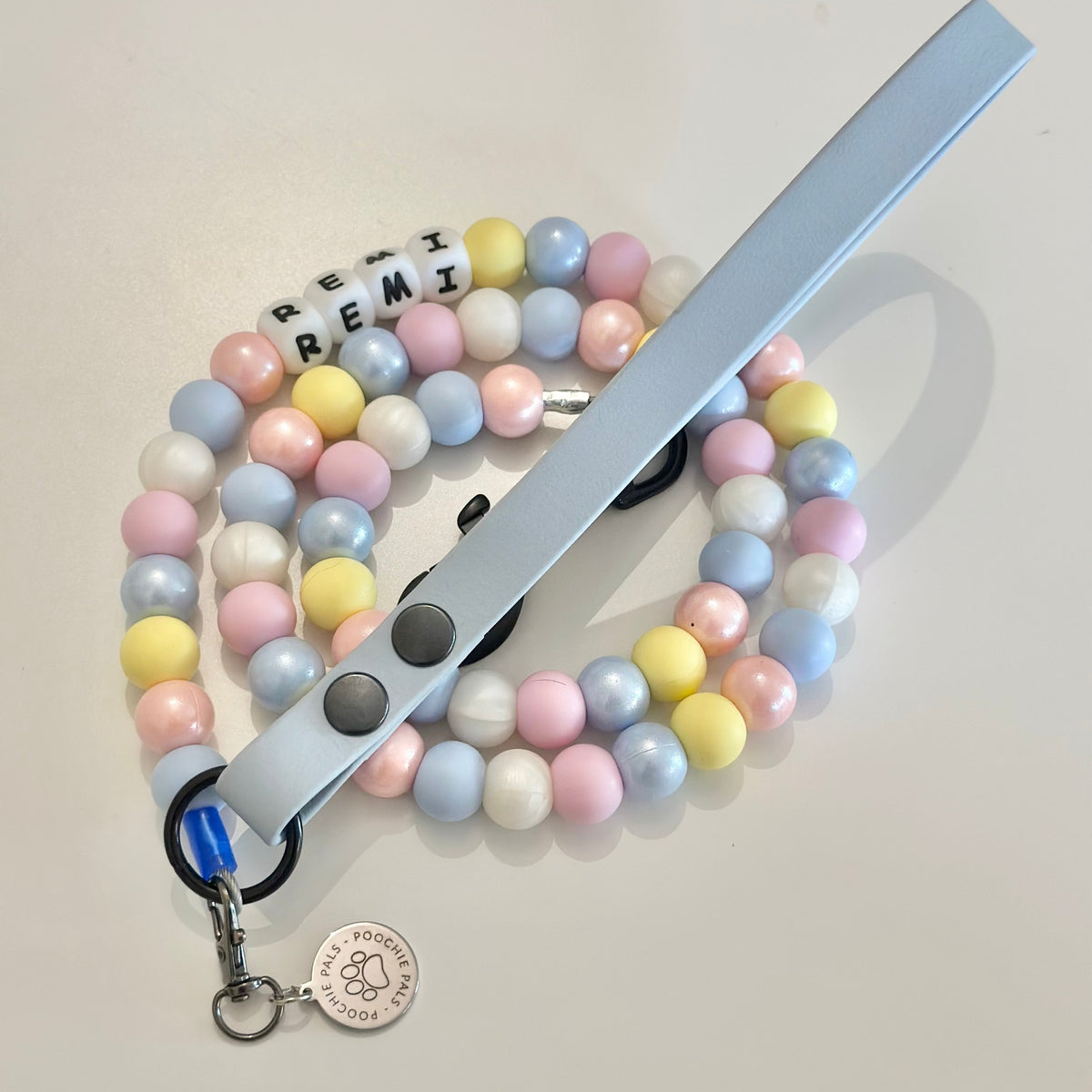 A pastel beaded pet leash with a light blue strap, featuring pastel-colored round beads in pink, yellow, blue, and white, a black snap button, and a silver clasp with a round brand tag.