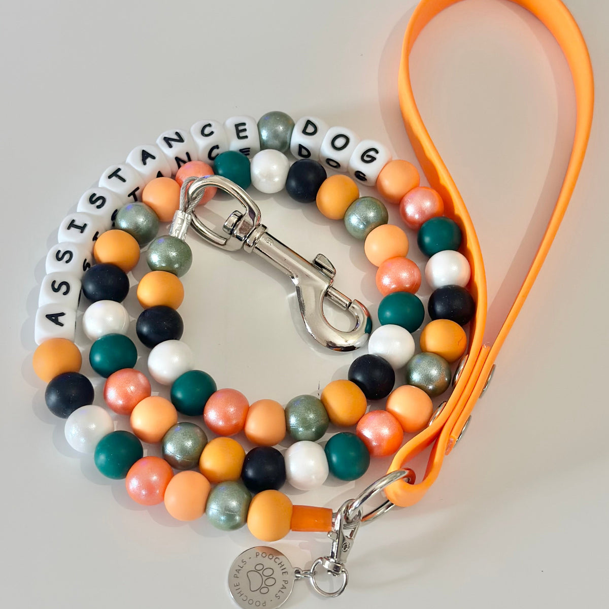 A beaded dog lead with an orange biothane handle, decorated with colourful round beads in orange, teal, green, white, and black, featuring white letter beads spelling 'ASSISTANCE DOG' and a silver metal clasp.
