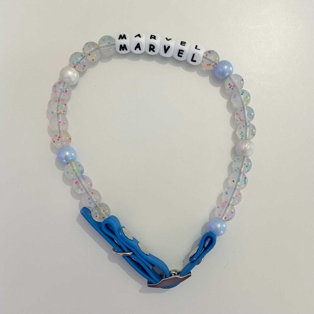 A beaded pet collar featuring translucent iridescent beads, white pearl-like beads, light blue beads, and white letter beads spelling 'MARVEL' twice. It has a bright blue collar strap with a silver metal clasp, photographed on a plain off-white background.